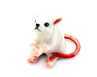 May include: A white ceramic figurine of a small animal, possibly a cat or dog, with a red tail and pink accents on its paws and ears. The figurine has black eyes and a black nose. The animal is sitting upright.