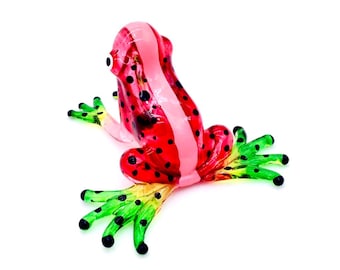 May include: A colorful glass frog figurine. The frog is primarily red with black spots and a pink stripe down its back. The feet are green with black tips, and the frog is set against a white background.