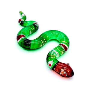 May include: A decorative glass snake in shades of green, red, and gold. The snake is coiled and has a detailed, textured design. The head is a deep red, transitioning to green with red and gold accents along the body.
