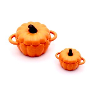 Può includere: Due pentole in ceramica arancione a forma di zucca con coperchi. La pentola più grande ha due manici e la pentola più piccola ha due manici.