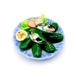 May include: Miniature plate with a meal of green mussels, sliced tomatoes, and a deviled egg. The mussels are open, revealing white and orange fillings. The plate is blue with a scalloped edge, and the food is arranged on a white background.