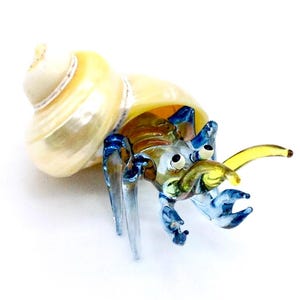Puede incluir: Figurita de cristal de un cangrejo ermitaño. El cangrejo tiene un cuerpo amarillo y marrón con patas y pinzas azules. Está dentro de una concha nacarada de color crema. Sobre un fondo blanco.