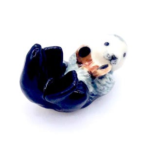 May include: A ceramic figurine of a sea otter lying on its back. The otter has a white face, gray body, and dark blue flippers. Its paws are a light brown color. The figurine is on a white background.