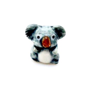 May include: A small ceramic koala figurine. The koala is mainly grey with black details, a white belly, and a reddish-brown nose. It has large, round ears and is seated.