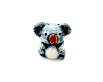 Może przedstawiać: Mała ceramiczna figurka koali. Koala jest głównie szary z czarnymi akcentami, białym brzuchem i czerwonobrązowym nosem. Ma duże, okrągłe uszy i siedzi.