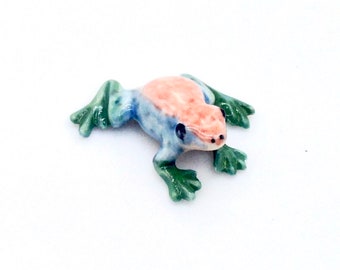 Op de afbeelding: Een klein keramiek kikker beeldje met een blauwe, groene en roze glazuur. De kikker zit op een wit oppervlak.