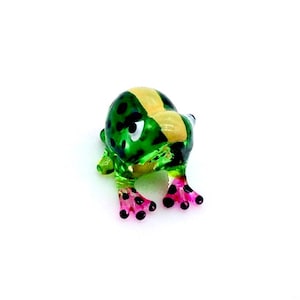 May include: A small glass frog figurine featuring a vibrant green body with black spots, a prominent yellow stripe running down the center of its back, and translucent pink feet with black tips, captured from a high-angle perspective.