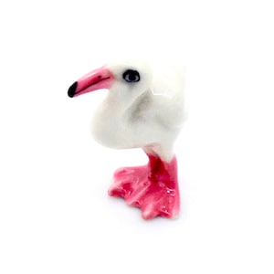 Pode incluir: Pequena figura de cerâmica de um pássaro branco com bico e pés rosa. O pássaro tem um olho preto e está sobre uma superfície branca. Um artigo decorativo.