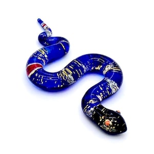 May include: A handcrafted glass snake figurine in a winding pose. The snake is primarily blue with silver and gold accents. A red stripe is visible on the body. The head is black with small pink details. The figurine is on a white background.