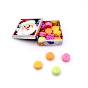 May include: A small, open box with a Santa Claus illustration on the lid, filled with colorful, miniature cookies. Several cookies in shades of orange, pink, yellow, and green are scattered around the box on a white surface.