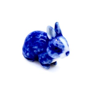 May include: A small, ceramic rabbit figurine in shades of blue and white. The bunny has a speckled pattern on its body and a white face and ears. The figurine is a decorative item, likely for display or collection.