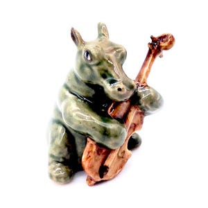 Könnte beinhalten: Eine Keramikfigur eines grünen Flusspferdes, das ein braunes Cello spielt. Das Flusspferd sitzt auf seinen Hinterbeinen und hält das Cello mit seinen Vorderbeinen.
