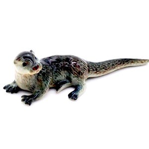 May include: A ceramic otter figurine in shades of gray, brown, and cream. The otter is lying down with its head and front paws raised. The figurine has a glossy finish and a red nose.