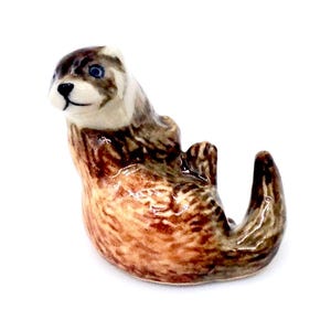 May include: A ceramic figurine of an otter in a playful pose. The otter has a brown and tan coat with blue eyes and a black nose. The figurine is detailed and appears to be hand-painted.