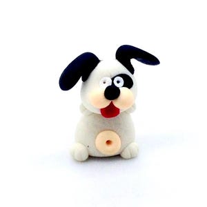 Könnte beinhalten: Eine kleine, weiße Hunde-Figur mit schwarzen Ohren und einem schwarzen Fleck über einem Auge. Der Hund hat eine rote Zunge, die herausragt, und einen beigen Bauchnabel. Die Figur sitzt auf einer weißen Oberfläche.
