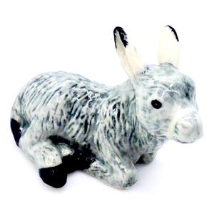 May include: A ceramic figurine of a donkey in a resting pose. The donkey is primarily gray with black accents on its ears and hooves. The figurine has a glossy finish and is set against a white background.