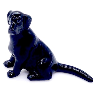 May include: A glossy black Labrador Retriever figurine in a seated position. The dog has a smooth, reflective surface and is set against a white background. The figurine's features are detailed, including its ears, paws, and tail.