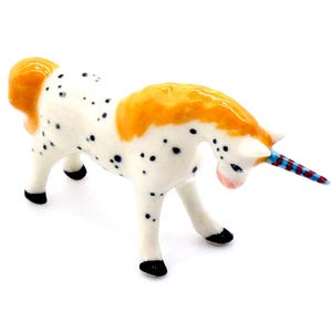 May include: A white ceramic unicorn figurine with black spots, a yellow mane and tail, and a blue and red striped horn.
