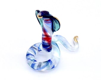 May include: A glass cobra figurine featuring a coiled body and an upright hood. The glass is clear with swirling patterns of red, white, and blue throughout the body, while the hood displays dark blue and teal tones with gold-coloured accents.