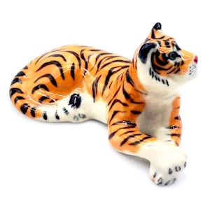 May include: A ceramic tiger figurine in a relaxed pose. The tiger has orange fur with black stripes, white underbelly and paws, and a red nose. The figurine is detailed and appears to be hand-painted.