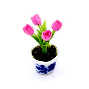 Könnte beinhalten: Eine Miniatur-Blumenarrangement aus rosa Blüten in einem weißen Keramiktopf mit blauen Blumendesigns. Der Topf ist mit brauner Erde gefüllt und die Blumen haben grüne Stiele.