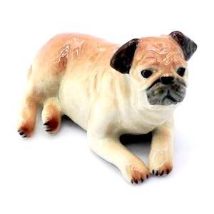 May include: A ceramic pug figurine in a lying down pose. The pug has a tan body with black accents on its ears, nose, and paws. The figurine is detailed with wrinkles and a realistic expression, set against a white background.