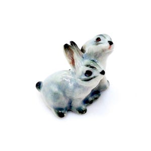 Op de afbeelding: Een kleine keramische figuur met twee lichtblauwe konijnen. De konijnen staan samen, de ene iets achter de andere. Het beeldje heeft zwarte ogen en oren met bruine punten. Gedetailleerd en delicaat ontwerp.