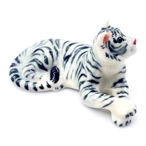 May include: A ceramic figurine of a reclining tiger. The tiger has a white base color with black stripes. The figurine has a pink nose and ear details. The tiger is lying down with its paws extended.
