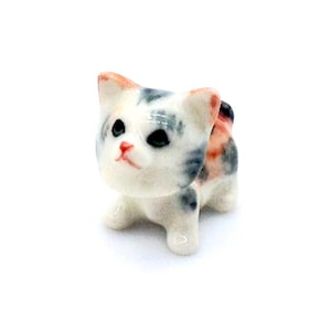 May include: A small, ceramic cat figurine in a standing pose. The cat has a white base color with patches of gray, orange, and black. It has green eyes and a pink nose and ears. The figurine is on a white background.