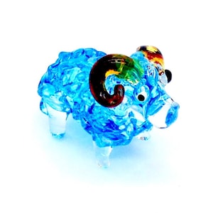 Op de afbeelding: Een klein blauw glazen beeldje van een ram met kleurrijke hoorns. De ram heeft een krullend, getextureerd lichaam en zwarte ogen.