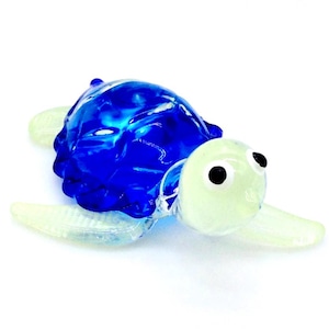 May include: A small glass sea turtle figurine with a blue shell and white body. The turtle has black eyes and a detailed shell.