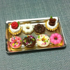以下が含まれることがあります： 銀色のトレイに、スプリンクルがかかったドーナツ、チョコレートドーナツ、白いフロスティングと赤と茶色のバラのついたカップケーキなど、7つのミニチュアデザートが載っています。