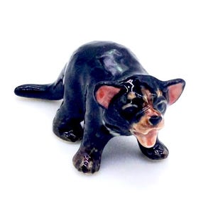 May include: A ceramic figurine of a Tasmanian devil. The figurine is primarily black with brown and pink accents on the face and ears. The devil is posed in a snarling stance, with its mouth open.