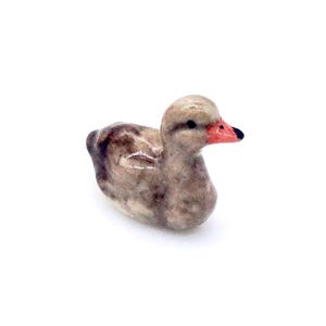 Peut inclure: Petite figurine de canard en céramique. Le canard a un corps brun clair avec des marques brun foncé. Son bec est rouge avec une pointe noire et de petits yeux noirs. La figurine est posée sur un fond blanc.