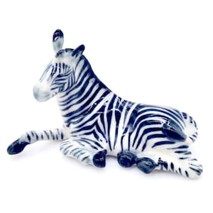 May include: A ceramic zebra figurine in a reclining pose. The zebra has a white base color with dark blue stripes. The figurine has a glossy finish and is set against a white background. The zebra's ears are upright, and its legs are extended.