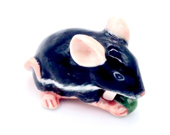 May include: A small ceramic figurine of a black mouse with pink ears and a white stripe on its back. The mouse is holding a green piece of food in its mouth.