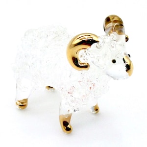 Peut inclure: Une figurine de bélier en verre blanc avec des cornes et des sabots peints en or. Le bélier est debout sur un fond blanc.
