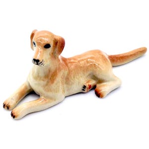 May include: A ceramic figurine of a golden Labrador Retriever dog in a relaxed, lying-down pose. The dog has a glossy finish, with black eyes and nose, and detailed paws. The figurine is a light tan color.