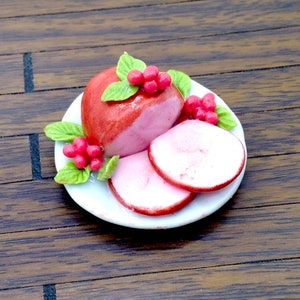 以下が含まれることがあります： 小さな白い皿に乗ったミニチュアの食品フィギュアで、2枚のスライスが添えられた艶出しされたハムのローストが特徴です。ハムは鮮やかな緑色の葉と小さな赤い実の房で飾られており、精巧でリアルな食品ディスプレイを作り出しています。