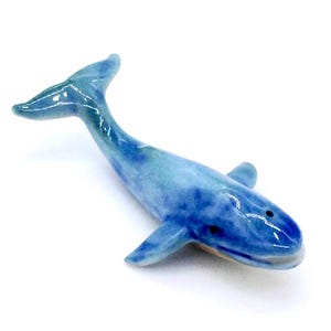 May include: A ceramic whale figurine in shades of blue. The whale has a glossy finish and is posed with its tail raised. The figurine has small fins and two small holes on the top of its head.
