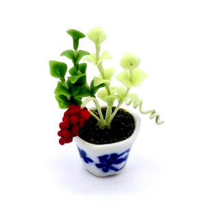 May include: Miniature ceramic potted plant with green and light green leaves, red berries, and a blue and white floral design on the pot. The plant is set against a white background.