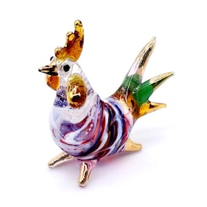 May include: A colourful glass rooster figurine with gold accents. The rooster features a white body with swirls of red, purple, and blue. The comb and tail feathers are gold, and the tail has a touch of green.