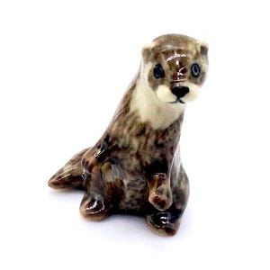 May include: A ceramic otter figurine in shades of brown, gray, and cream. The otter is depicted in a seated position, with its front paws raised. The figurine has a glossy finish and detailed features.