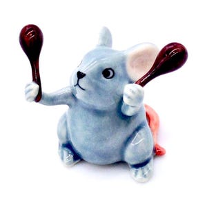 May include: A small, blue ceramic figurine of a mouse holding two red maracas. The mouse has a pink tail and is sitting on its hind legs.