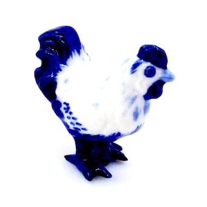 May include: A ceramic rooster figurine, mainly white with blue detailing. The comb, tail, and feet are blue. The figurine is set against a white background. The rooster is a decorative item.