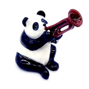 Puede incluir: Una figurita de panda blanco y negro tocando una trompeta roja. El panda está sentado en sus ancas con las patas delanteras levantadas hacia la trompeta. La figurita está hecha de cerámica y tiene un acabado brillante.