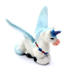 May include: A ceramic figurine of a winged unicorn, featuring a white body, blue wings, and a blue and red striped horn. The unicorn has a pink muzzle and a decorative necklace with red and blue accents. The figurine is posed in a seated position.