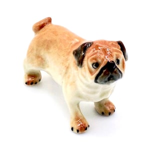 May include: A ceramic pug figurine in a standing pose. The pug has a tan body with black accents on its face, ears, and paws. The figurine is detailed with realistic features and a glossy finish. The pug is a popular breed.