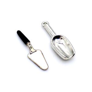 May include: Two miniature silver tools: a small trowel with a black handle and a scoop with a rounded edge.