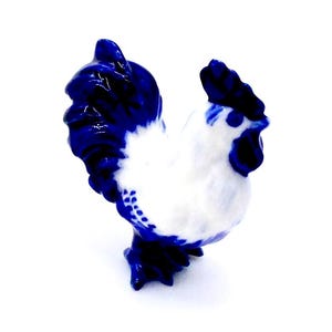 May include: A white and blue ceramic rooster figurine. The rooster has a blue comb and wattle, and its tail feathers are also blue. The figurine is a traditional folk art style.